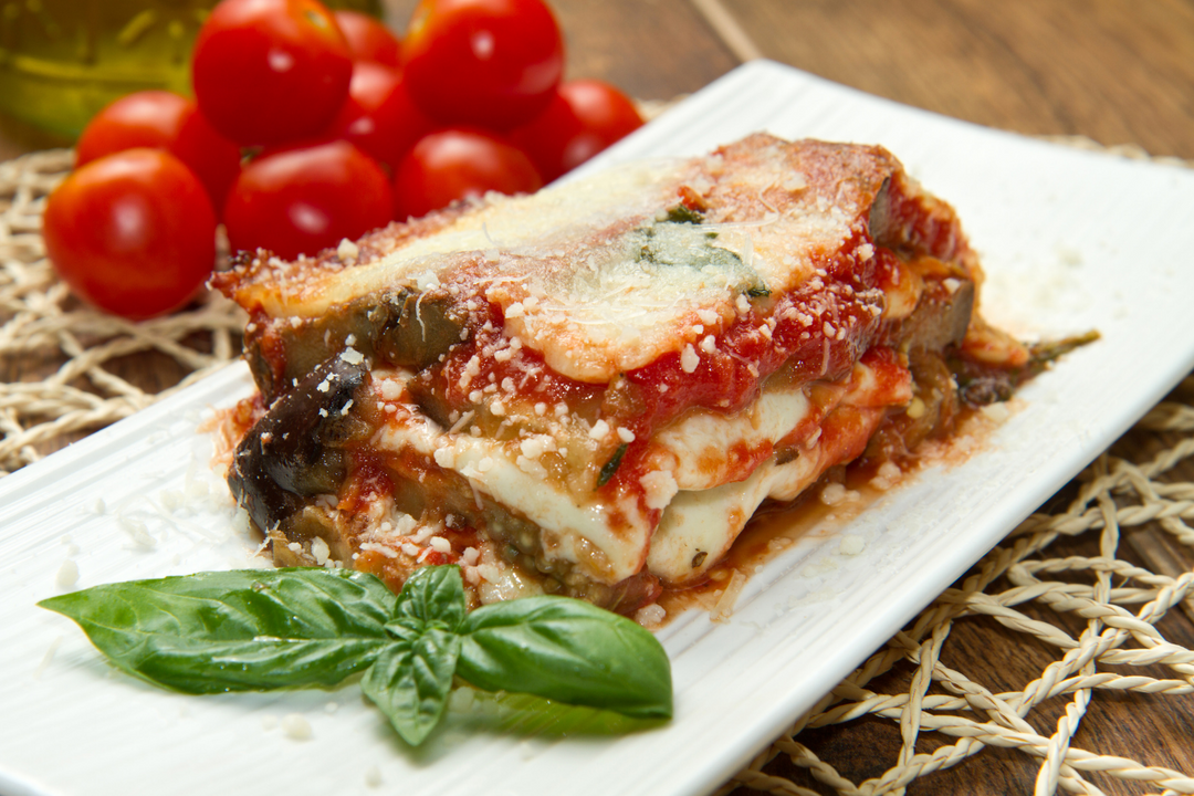 parmigiana di melanzane servita su piatto bianco con sfondo pomodori freschi